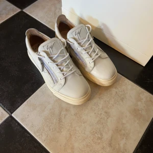 Vita sneakers från Giuseppe Zanotti - Säljer ett par vita sneakers från Giuseppe Zanotti med snörning och dragkedja på sidorna. Skorna har en platt sula och rund tå, samt märkets logga på plösen. Perfekta för en stilren och trendig look.