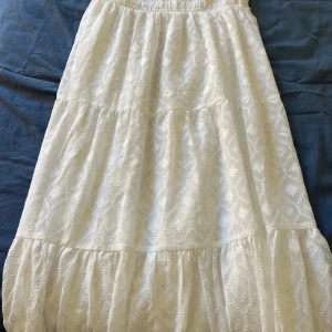 Vit mönstrad långkjol från Gina Tricot - Säljer en vit långkjol från Gina Tricot med diskret geometriskt mönster och resår i midjan. Kjolen har flera volanger och är luftig, perfekt för varmare dagar. Aldrig använd!