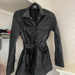 Svart skinnjacka med bälte från Gina Tricot - Säljer en svart skinnjacka från Gina Tricot med bälte i midjan och stora fickor framtill. Jackan har långa ärmar, klassisk krage och knappar framtill. Perfekt för dig som vill ha en edgy look.