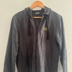 Mörkgrå hoodie från EA7 - Säljer en mörkgrå hoodie från EA7 med dragkedja och huva. Tröjan har svarta snören och EA7-logga i gult på bröstet. Perfekt för en avslappnad stil och enkel att matcha med det mesta.