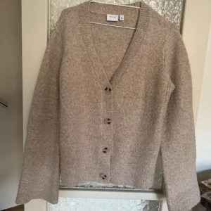 Beige stickad kofta från VILA - Mysig beige stickad kofta från VILA med v-ringning och knappar framtill. Koftan har långa ärmar och bred ribbad kant vid ärmslut och nederkant. Perfekt för lager-på-lager nu till sommar och vår.