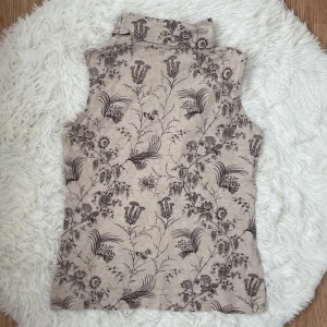 Beige blommig topp  - Säljer en beige väst med svart blommigt mönster. Västen har hög krage och är ärmlös, perfekt för att addera lite extra stil till din outfit. 