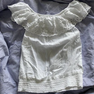 Vit blus med volang och broderade detaljer - Säljer en söt vit blus med bred volangkrage och broderade blommönster. Blusen har resår nertill och är perfekt för sommardagar. Den är ärmlös och har en romantisk känsla med sina fina detaljer. Storlek 158 men skulle säga att den passar xxs/xs 🫶🏻