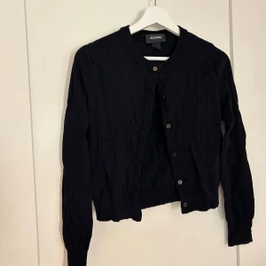 Svart kofta från Monki - En klassisk svart kofta från Monki med rund halsringning och knäppning framtill. Tröjan har långa ärmar och är perfekt att slänga över en topp eller klänning. Enkel och stilren design som passar till det mesta.