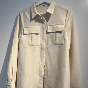 Beige skjorta med fickor från NA-KD - Stilren beige skjorta från NA-KD med två stora bröstfickor och klassisk krage. Skjortan har långa ärmar och knäppning framtill. Perfekt att styla lager på lager eller bära som den är.