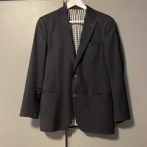 Mörkblå kavaj från Oscar Jacobson - Mörkblå kavaj från Oscar Jacobson med två knappar framtill. Fodret har ett rutigt mönster i vitt och blått, samt beige inuti armarna med fina detaljer (bild 4). Perfekt till skolavslutning, bal eller fest, skick 10/10, säljer pga att den är för liten för mig. Nypris: ca 5000kr 
