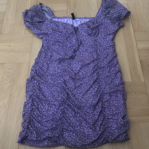 Lila blommig kortklänning - Supersöt lila kortklänning med småblommigt mönster och lätt rynkade detaljer. Klänningen har korta puffärmar och knytning fram. Bild på kan skickas privat :) 