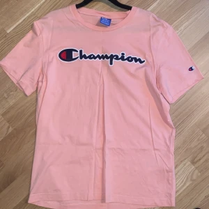 Rosa t-shirt från Champion - Säljer en rosa t-shirt från Champion med klassisk logga broderad på bröstet och liten logga på ärmen. T-shirten har normal passform och är tillverkad i mjuk bomull. Perfekt för en avslappnad och sportig stil.