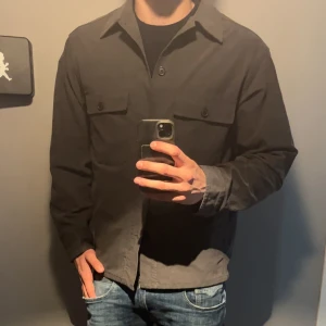 overshirt med knappar - Säljer en stilren svart overshirt med klassisk krage och knäppning framtill. Två bröstfickor med lock och knappar ger en clean look. Perfekt att bära över en t-shirt eller hoodie.