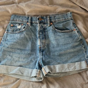 Blå jeansshorts från 157 - Klassiska blå jeansshorts från 157 med fem fickor och uppvikta benslut. De har normal passform och stängs med fyra knappar. Perfekta för sommaren och enkla att matcha med olika toppar.