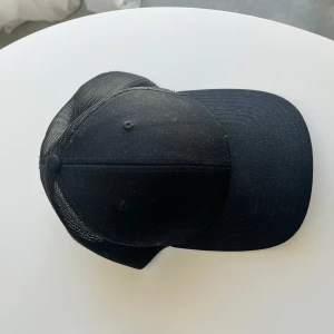 Svart truckerkeps från Classic - Säljer en svart truckerkeps från Classic med böjd skärm och justerbar snapback. Kepsen har mesh på baksidan.