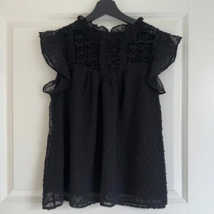 Svart blus med spets och volang - Svart blus med genombruten spets upptill och volangdetaljer vid ärmhålen. Blusen är ärmlös och har en luftig, lätt transparent känsla med diskret prickigt mönster i tyget. Perfekt för en trendig och elegant look.