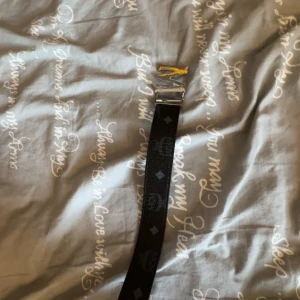 Mcm bälte - Säljer ett svart bälte från MCM med silverfärgat MI-spänne. Bältet har diskreta mönster i form av små rutor och loggor längs hela bandet. Perfekt accessoar för att lyfta din outfit.