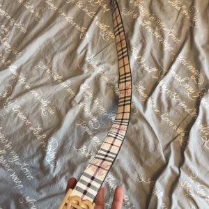 Burberry bälte  - Snyggt bälte med klassiskt rutigt mönster i beige, svart, rött och vitt. Bältet har ett stilrent guldigt spänne och passar perfekt för att lyfta en outfit. Tidlös design som funkar till många stilar.