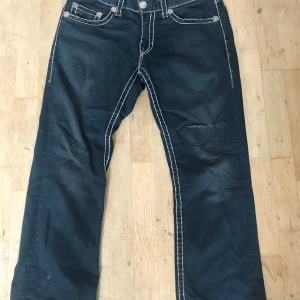 True Religion jeans med vita sömmar - Säljer ett par mörkblå True Religion jeans med tydliga vita kontrastsömmar och klassiska bakfickor med broderad logga. Jeansen har normal passform och raka ben, perfekt för en avslappnad stil.