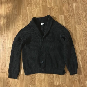 Svart stickad kofta från John Henric - Säljer en svart stickad kofta från John Henric i storlek L. Koftan har ribbad struktur, knäppning framtill med fem knappar och en bred sjalkrage. Perfekt för lager-på-lager och passar till många olika stilar.