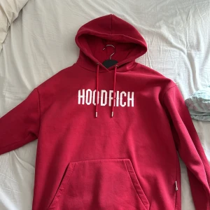 Röd hoodie från Hoodrich - Säljer en röd hoodie från Hoodrich med vit logga tryckt på bröstet. Tröjan har huva med dragsko och en stor magficka framtill. Perfekt för dig som gillar streetwear och vill ha en snygg och bekväm tröja.