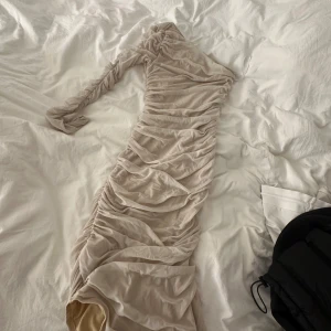 Beige långklänning med rynk och oneshoulder - Säljer en beige långklänning med rynkade detaljer och oneshoulder-design. Klänningen har en lång ärm och är figurnära med snygg drapering längs hela plagget. Perfekt för dig som vill sticka ut med en unik och trendig look.