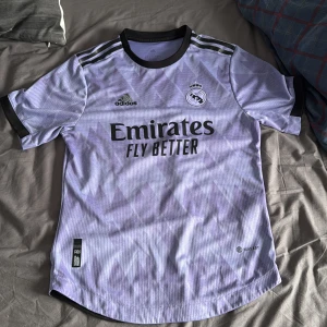Real Madrid bortatröja 2022/23 - Säljer en ljuslila Real Madrid bortatröja från Adidas med svarta detaljer och klubbmärke på bröstet. Tröjan har korta ärmar, svart krage och subtilt mönster på tyget. Perfekt för fotbollsfans som vill visa sitt stöd. Det står M på den men den är S