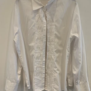 Vit skjorta med veckad front - Klassisk vit skjorta med lång ärm och snygga veckade detaljer framtill. Skjortan har krage och knäppning framtill, perfekt för en stilren look. Tillverkad i bomull för en skön känsla och tidlös stil.