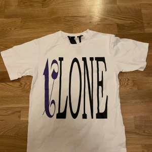 Vit t-shirt från VLONE x Palm Angels - Vit t-shirt med stor svart och lila VLONE-logga framtill och stor lila V med Palm Angels-tryck på ryggen. Klassisk passform och rund hals. Perfekt statement-plagg för streetwear-stilen.