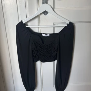 Svart croppad topp från Sisters Point - Trendig svart croppad topp från Sisters Point med långa puffärmar och snygg rynkdetalj framtill. Offshoulder-modell som ger en cool vibe och passar perfekt till höga jeans eller kjol. Mjuk och stretchig polyester för en bekväm känsla.