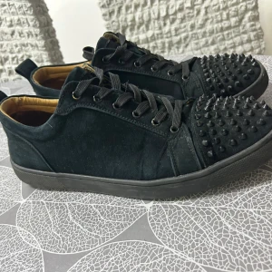 Christian Louboutin svarta sneakers med nitar storlek 47 - Exklusiva svarta sneakers från Christian Louboutin i mocka med coola svarta nitar på tån och klassisk röd sula. Skorna har snörning och en låg profil, perfekt för dig som vill sticka ut med en edgy och lyxig stil.