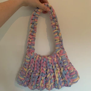Pastellfärgad virkad axelväska - Superfin axelväska i chunky virkat material med breda flätor. Väskan har pastelliga färger i lila, rosa, blått och gult och en mjuk, handgjord känsla. Perfekt storlek för det viktigaste och ger en färgklick till din outfit.
