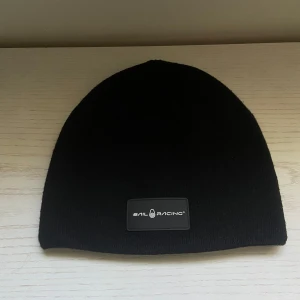 Svart mössa från Sail Racing - En stilren svart mössa från Sail Racing med en diskret logotyp framtill. Mössan är enkel i designen och tillverkad i mjukt stickat material som sitter skönt på huvudet. Perfekt för kalla dagar när du vill hålla värmen och ändå se clean ut.