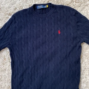 Mörkblå kabelstickad tröja Polo Ralph Lauren - Kabelstickad mörkblå tröja från Polo Ralph Lauren med klassisk rund halsringning och röd broderad logga på bröstet. Perfekt för lager-på-lager under kyliga dagar. Priset kan diskuteras vid snabbt köp! 