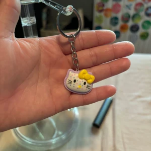 Hello Kitty nyckelring med gul rosett - Söt Hello Kitty nyckelring med ett vitt ansikte, lila kant och en stor gul rosett. Ansiktet har färgglada prickar och klassiska svarta morrhår. Perfekt accessoar för att piffa upp dina nycklar eller väska.