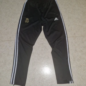 Real Madrid Adidas svarta träningsbyxor - Äkta från Adidas! Svarta träningsbyxor från Adidas med Real Madrid-logga i guld och klassiska vita ränder längs sidorna. Byxorna har smal passform, resår i midjan och är gjorda i polyester. Perfekta för fotbollsträning eller chill.