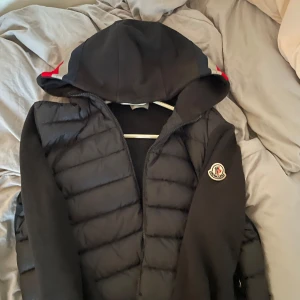 Svart pufferjacka från Moncler - Svart cardigan från Moncler med huva och coola röda och grå detaljer på huvan. Jackan har quiltad front, dragkedja och Moncler-logga på ärmen. Perfekt för hösten och våren. Säljer till ett lägre pris då den har en liten defekt på armen samt att den är lite solblekt. 