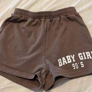 Bruna shorts från SHEIN med tryck - Mysiga bruna shorts från SHEIN med elastisk midja och vit text 'BABY GIRL 90'S' på ena benet. Perfekta för chill dagar eller träning. Mjukt material och loose fit för maximal komfort.