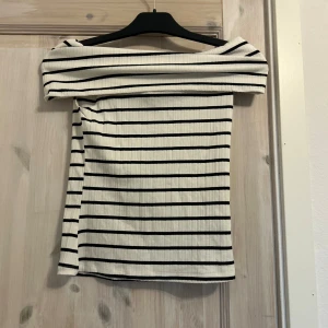 Randig offshoulder topp i ribbad bomull - Säljer en vit och svart randig offshoulder topp i ribbad bomull. Toppen har en snygg omlottkänsla över axlarna och är figurnära med korta ärmar. Perfekt för dig som gillar en stilren och trendig look.