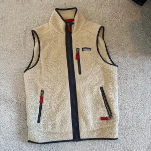 Beige fleeceväst från Patagonia - Säljer en beige fleeceväst från Patagonia med marinblå detaljer och röda dragkedjor. Västen har hög krage, tre fickor med dragkedja och är perfekt för lager-på-lager. Klassisk och stilren design som funkar året runt.