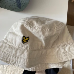 Beige bucket hat från Lyle & Scott - Stilren beige bucket hat från Lyle & Scott med klassisk gul broderad logga framtill. Den har en avslappnad passform som gör att den passar många Perfekt accessoar för soliga dagar och enkel att matcha med olika outfits🥰🥰