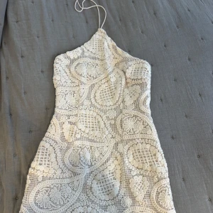 Beige virkad klänning från PrettyLittleThing - Supersöt kort klänning, beige virkad, från PrettyLittleThing. Halterneck med knyt i nacken och figurnära passform. Klänningen har ett unikt virkat mönster med blommiga detaljer och är fodrad. Perfekt för sommarens alla tillfällen!