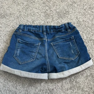 Blå jeansshorts från Lindex - Snygga blå jeansshorts från Lindex med klassisk femficksdesign, uppvikta benslut och gylf med knapp. Shortsen har normal passform och är perfekta för varma dagar. Elastisk midja för extra komfort.