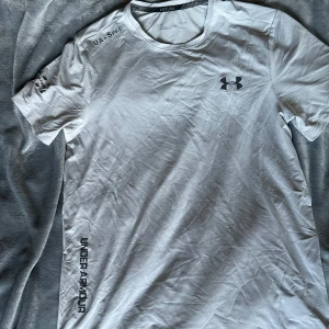 Grå träningst-shirt från Under Armour - Snygg vit t-shirt från Under Armour med svart logga på bröstet och tryck på ärmen och sidan. Tillverkad i ett lätt och andningsbart material som passar perfekt för träning. Klassisk rund hals och korta ärmar för en sportig look.