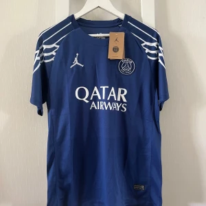 PSG x Jordan blå fotbollströja M - Snygg blå Paris Saint-Germain fotbollströja från Jordan med vita detaljer och loggor. Tröjan har korta ärmar, rund hals och coola vita mönster på axlarna. Materialet är lätt och andas, perfekt för match eller träning. Qatar Airways tryck på bröstet.