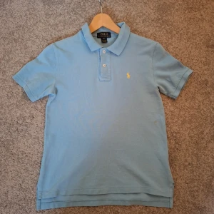 Ljusblå pikétröja från Ralph Lauren - Nypris 700 kr.  Snygg ljusblå pikétröja från Ralph Lauren med klassisk krage och två knappar framtill. Ikoniska gula loggan broderad på bröstet. Tillverkad i mjuk bomull och har korta ärmar. Storlek M (För barn 11-12)