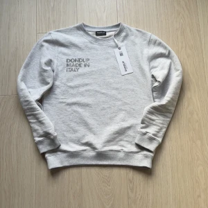 Ljusgrå sweatshirt från Dondup - Snygg ljusgrå sweatshirt från Dondup med broderad text 'Dondup Made in Italy' på bröstet. Tröjan har rund hals, ribbade muddar och är tillverkad i mjuk bomull. Perfekt för en chill och stilren look. Helt ny aldrig använd, kvitto finns 