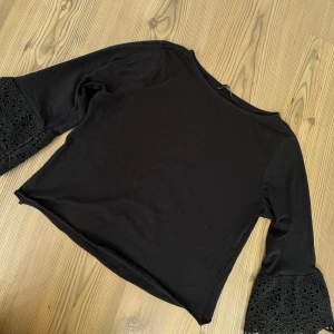 Svart blus med spetsärmar - Snygg svart blus med trekvartsärmar och breda spetsdetaljer längst ner på ärmarna. Blusen har en rak passform och är tillverkad i mjuk bomull. Perfekt för dig som gillar stilrena plagg med en twist!