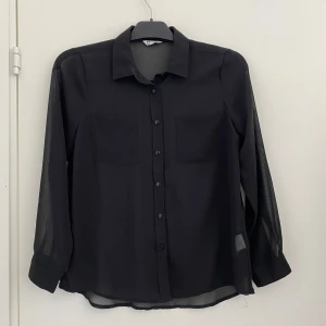 Svart transparent blus med knappar, stl 152 - Stilren svart skjorta med transparent känsla, klassisk krage och två bröstfickor. Långa ärmar med manschett och knappar framtill. Perfekt för dig som gillar en enkel men snygg look. Passar till många olika outfits.                                                                             Hör av dig vid frågor!                                            Köparen står för frakten. 