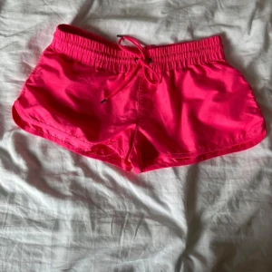 Neonrosa shorts med snörning - Snygga neonrosa shorts med elastisk midja och snörning framtill. Modellen är loose fit med rundade benslut och tillverkad i ett lätt syntetmaterial som passar perfekt för sommarens aktiviteter eller träning. Riktigt poppig färg som sticker ut