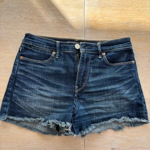 Mörkblå jeansshorts Abercrombie - Snygga mörkblå jeansshorts från Abercrombie & Fitch med fransig kant nertill och klassisk femficksdesign. Shortsen har normal passform, dragkedja och knapp framtill samt slitna detaljer för en chill vibe. Perfekta för varma dagar och enkel att matcha.