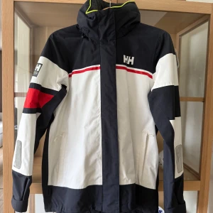 Helly Hansen vindjacka i svart och vitt - Snygg vindjacka från Helly Hansen i svart, vitt och rött med coola färgblock och HH-logga på bröstet. Jackan har huva, dragkedja och praktiska fickor. Perfekt för blåsiga dagar och till havs.  Regular fit och sportig look.