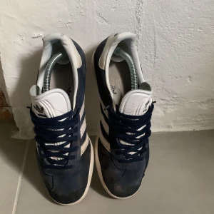 Adidas Gazelle mörkblå sneakers mocka - Snygga Adidas Gazelle sneakers i mörkblå mocka med klassiska vita ränder och vit plös med Adidas-logga. Skorna har rund tå, platt sula och svarta snören. Perfekta för dig som gillar retrostil och streetwear.