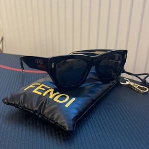 Svarta solglasögon från Fendi - Snygga svarta solglasögon från Fendi med breda skalmar och guldiga detaljer. Fendi-logga på sidan och coolt mönstrad metallinsats på insidan av skalmen. Kommer med original svart fodral med gul Fendi-text. Dem är 100% äkta (synlig kvalitet) och är i topp skick med inga skador. Köpta för runt 3000